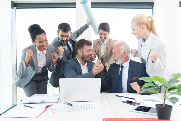 Two businessmen doing a victory clasp while the colleagues happily cheer around